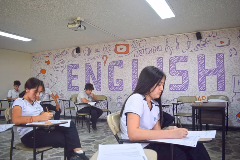 Estudiantes escriben en un aula decorada con un gran mural de la palabra «English».