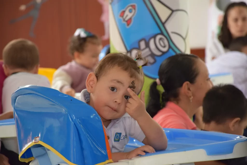 Estudiante de preescolar con la mano en la frente, en una silla alta azul, junto a otros niños del aula.
