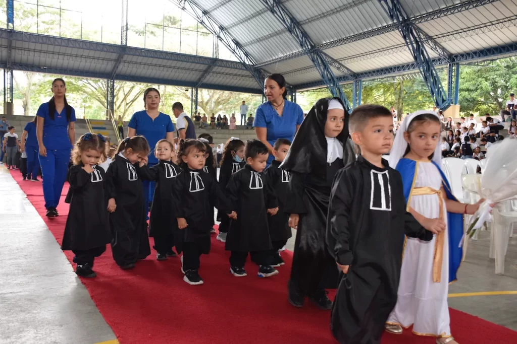Niños de preescolar con túnicas negras desfilan por una alfombra roja en el coliseo, acompañados por sus docentes.