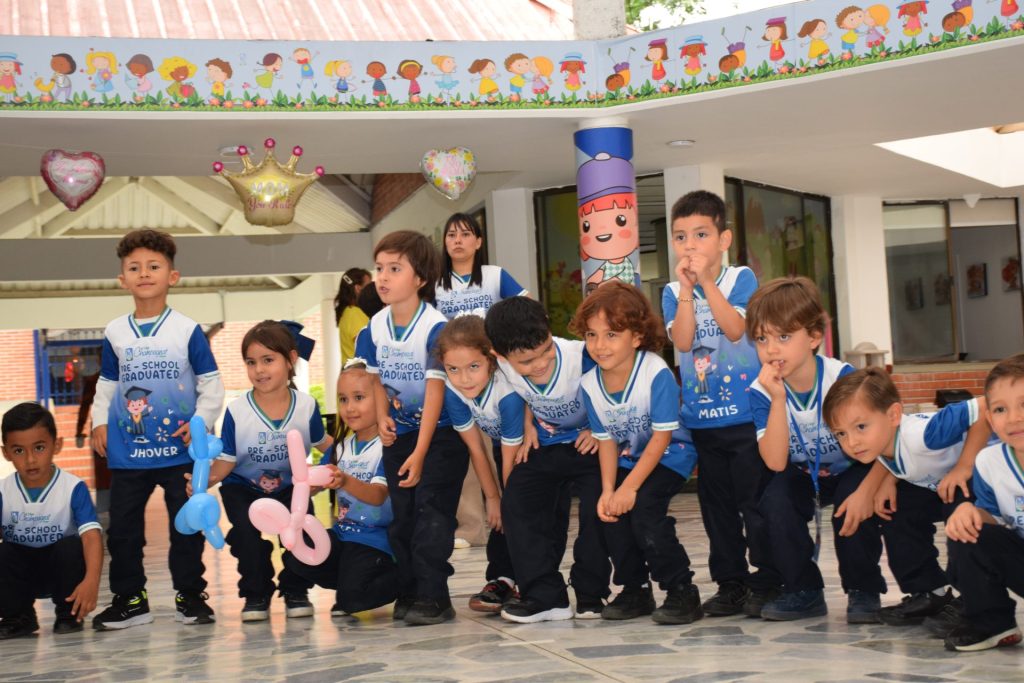 Niños de preescolar con camisetas de graduación posan en un patio del colegio decorado con globos.