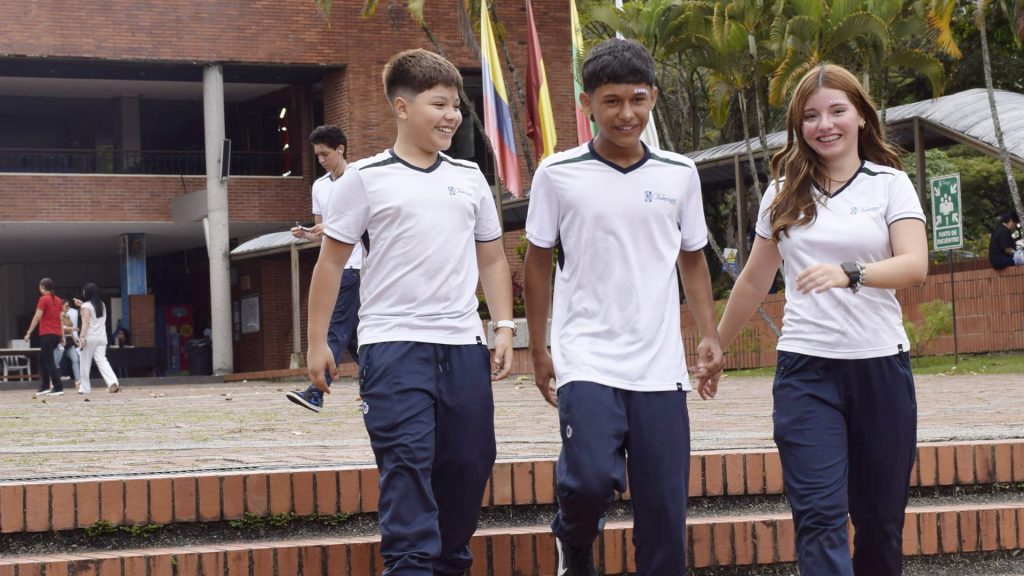 Tres estudiantes de bachillerato caminan sonrientes por la plazoleta del colegio.